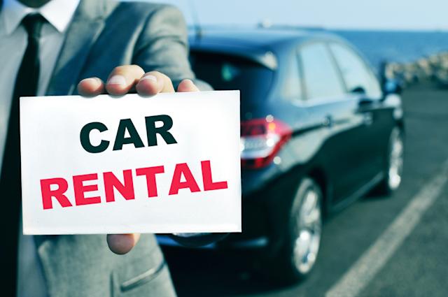 Person holding 'Car Rental' sign near a parked car by the sea.